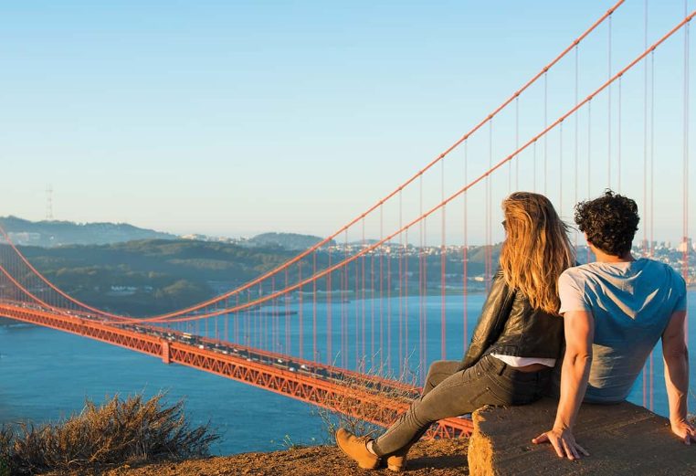 A man and woman sit on a rock up on a hill, staring out over the Golden Gate Bridge in San Francisco, California.