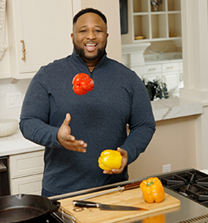 Chef Jernard Wells stands in front of a kitchen stovetop, smiling while holding a yellow bell pepper and throwing a red bell pepper in the air.