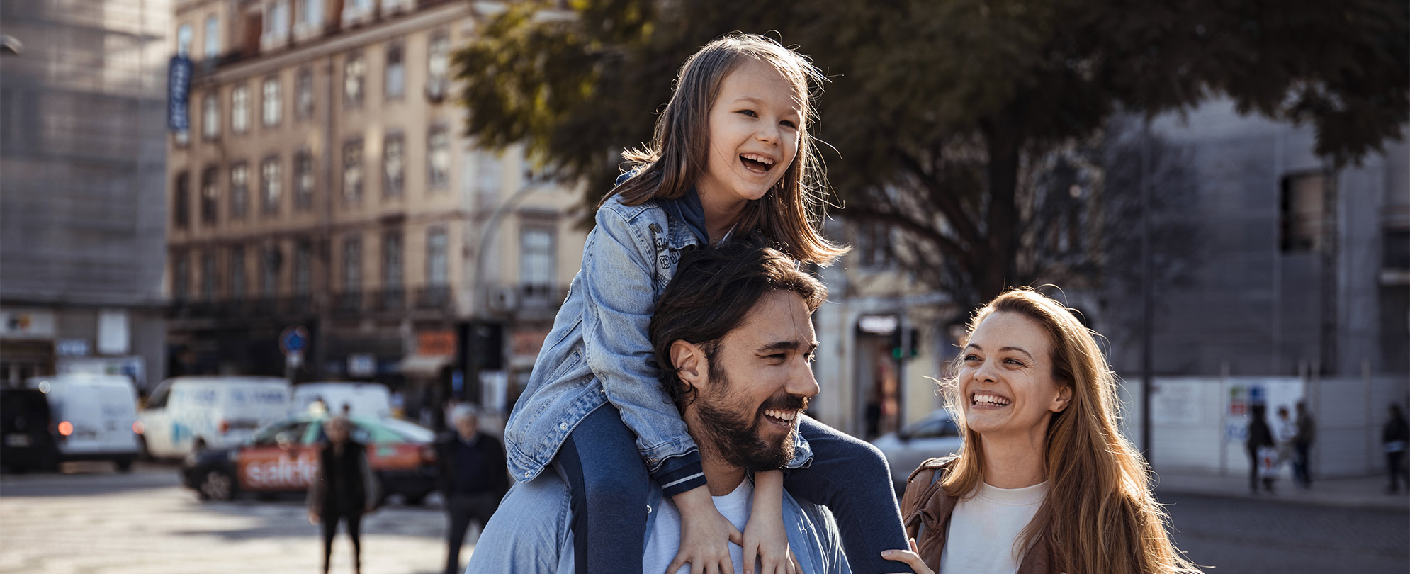 A smiling dad carries his child on his shoulders while mom smiles at them.