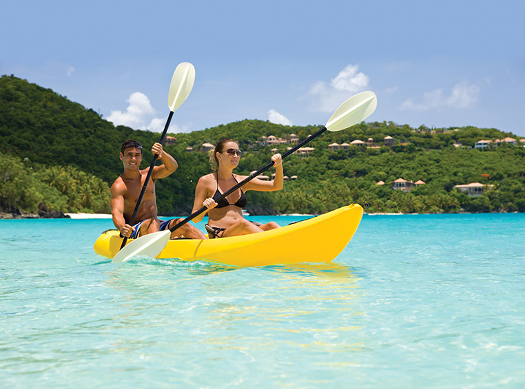 A couple paddling in a yellow kayak across clear blue waters. 