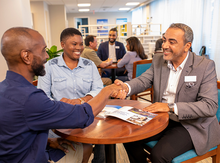 A WorldMark by Wyndham associate sits at a table with a smiling man and woman, he shakes the man’s hand.