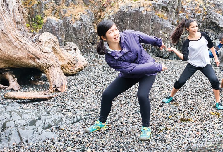 A mom, dad, and teenage daughter stand on a rocky shoreline focus as they throw stones into the water.