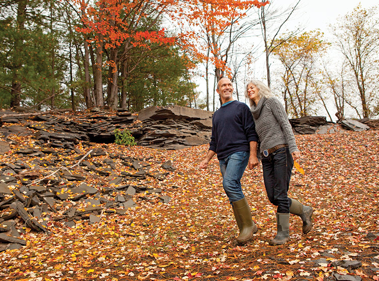 An older man and woman holding hands walk down a trail covered in fall leaves.