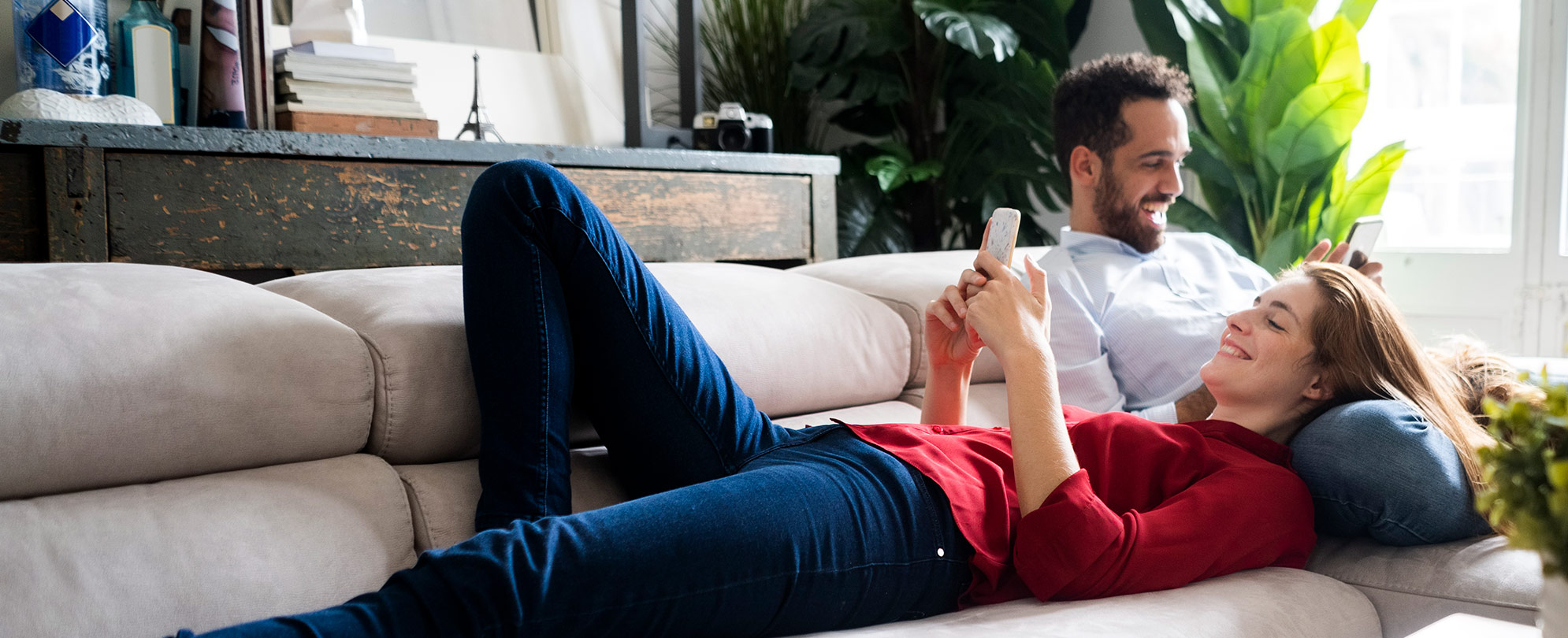 A smiling man and woman laying on a sofa while looking at their phones in a modern looking room.