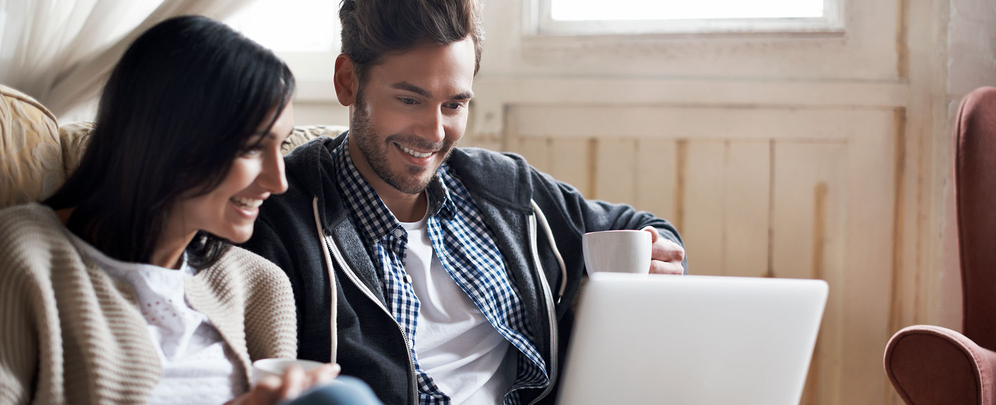 A young man and woman holding coffee mugs smile down at a laptop.