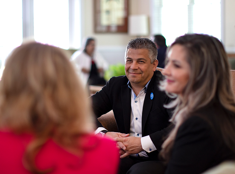 Two people are seated at a table looking and smiling at the person speaking at the head of the table.