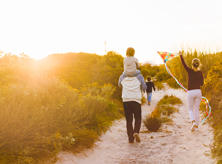 A family walks along a sandy trail, the dad carries their young child on his shoulders while the mom flies a kite.