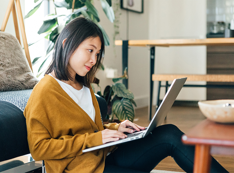 A woman sits on the floor in front of her couch, smiling at her laptop.