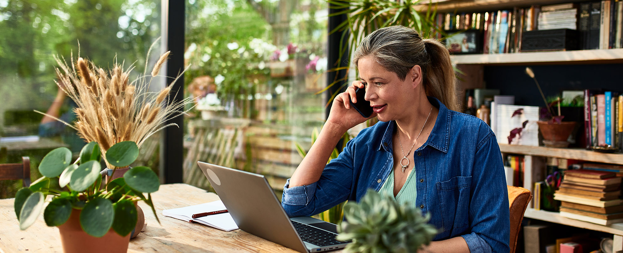 A woman sits at a desk in her home office and looks at her laptop as she speaks to someone on her cell phone.