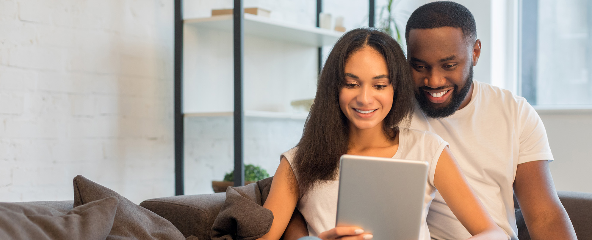 Happy young couple sitting on a couch, smiling down at a tablet. 