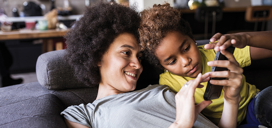 A mom and son lay on a couch looking at a cellphone together.