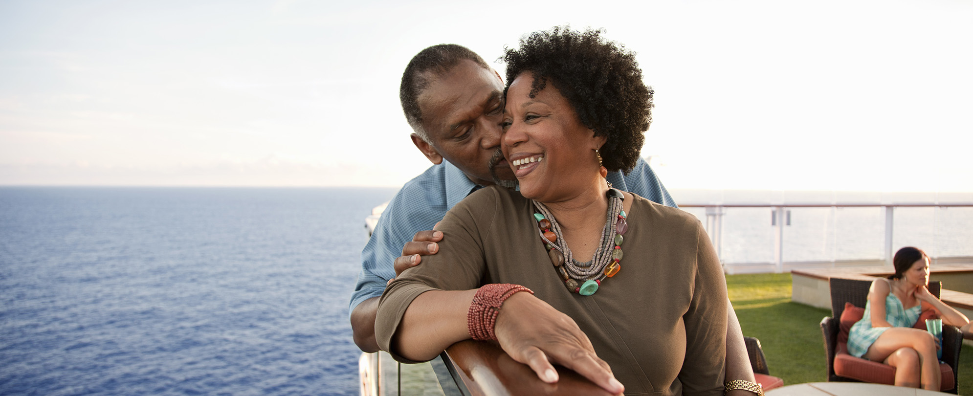 An older man kissing his wife's cheek while smiling on a deck overlooking the water.