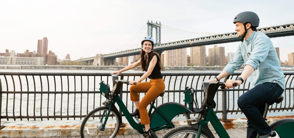 Happy couple bikes past a bridge in New York City, one of many Club Wyndham destinations available to WorldMark owners through Wyndham Club Pass.