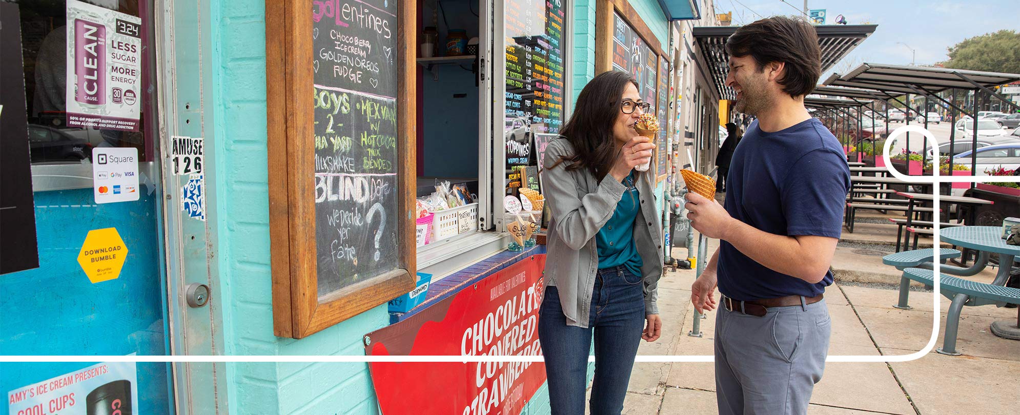 A smiling man and woman enjoy ice cream cones by the window of the ice cream storefront.