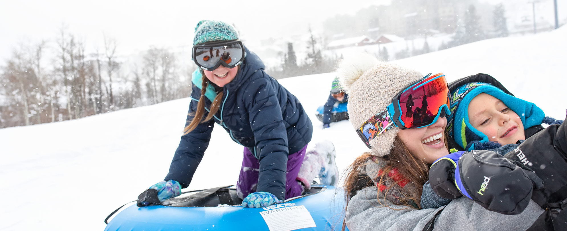 A smiling family in coats, hats, and ski goggles ride snow tubes during their WorldMark by Wyndham winter vacation.