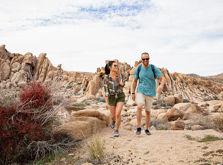 Smiling man and woman wearing sunglasses and backpacks hike along rocky path during their WorldMark by Wyndham vacation.